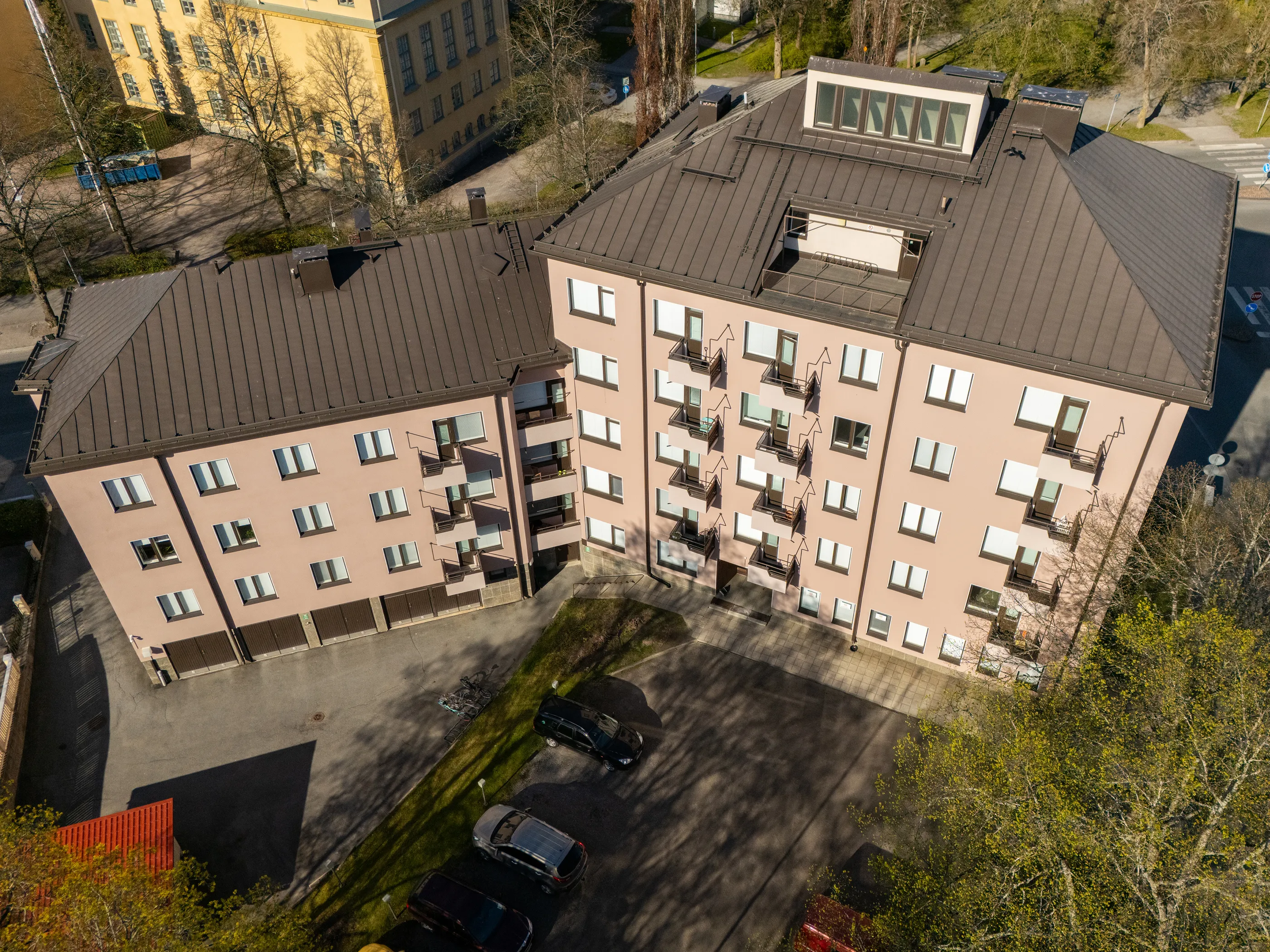 Ilmakuva kahdesta vaaleanpunaisesta asuinrakennuksesta, joissa on tummanruskeat peltikatot. Suuremmassa rakennuksessa on kattoikkunoita ja kattoterassi, pienemmässä useita parvekkeita. Rakennusten ympärillä on parkkipaikka autoineen ja polkupyörineen, vihreitä nurmialueita ja puita, joissa on sekä paljaita oksia että uusia vihreitä lehtiä. Kirkas auringonvalo luo pitkiä varjoja. Taustalla näkyy keltainen rakennus ja sininen jäteastia.