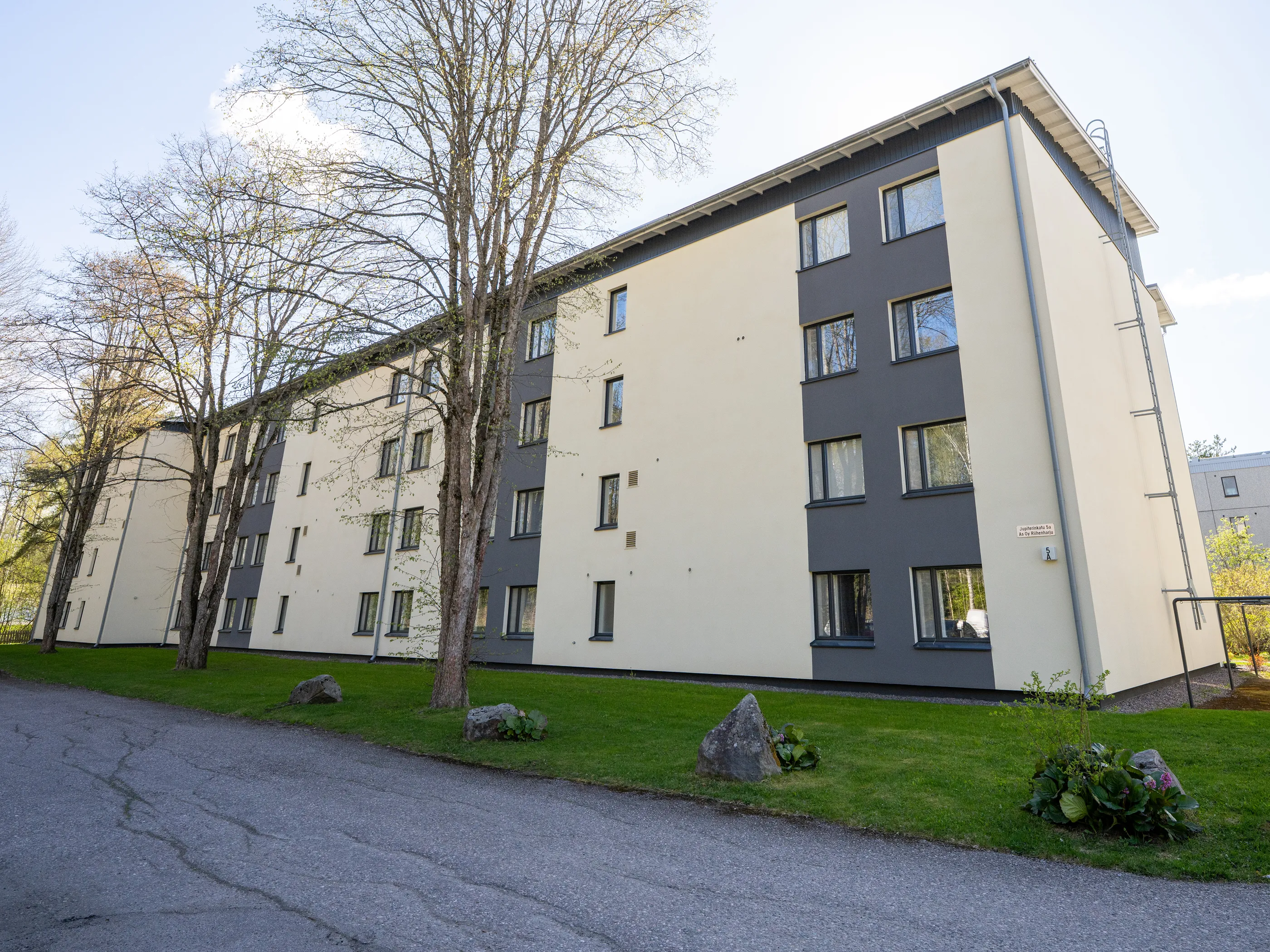 Pitkä, monikerroksinen asuinrakennus, jonka seinät ovat vuorotellen vaaleanbeiget ja tummanharmaat. Rakennuksessa on useita tummakehyksisiä ikkunoita, räystäskouru ja oikealla puolella palotikkaat. Rakennuksen edessä on vihreää nurmikkoa, useita puita, joissa on vähän lehtiä, sekä kiviä ja kasveja. Etualalla on asfalttitie. Kirkas päivänvalo valaisee kohteen sinisen taivaan ja pilvien alla. Rakennuksen seinässä on kyltti, jossa lukee "Jupiterintähti 5a As Oy Riimenharju" ja "5 A".